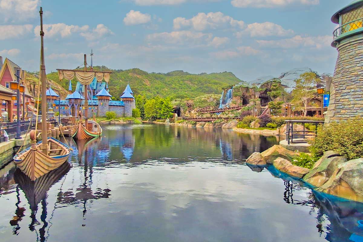 a large river with wooden ships going through what looks like a viking town with green forest in the back ground