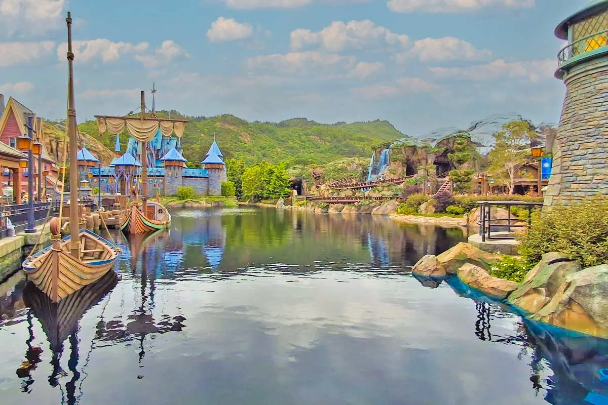 a large river with wooden ships going through what looks like a viking town with green forest in the back ground 