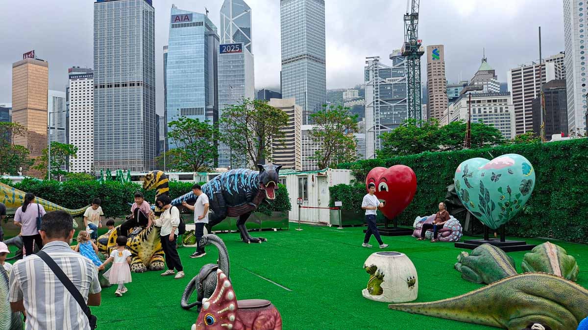 big dinosaur replicas on fake grass in a park below large skyscrapers
