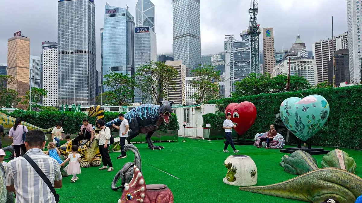 big dinosaur replicas on fake grass in a park below large skyscrapers 